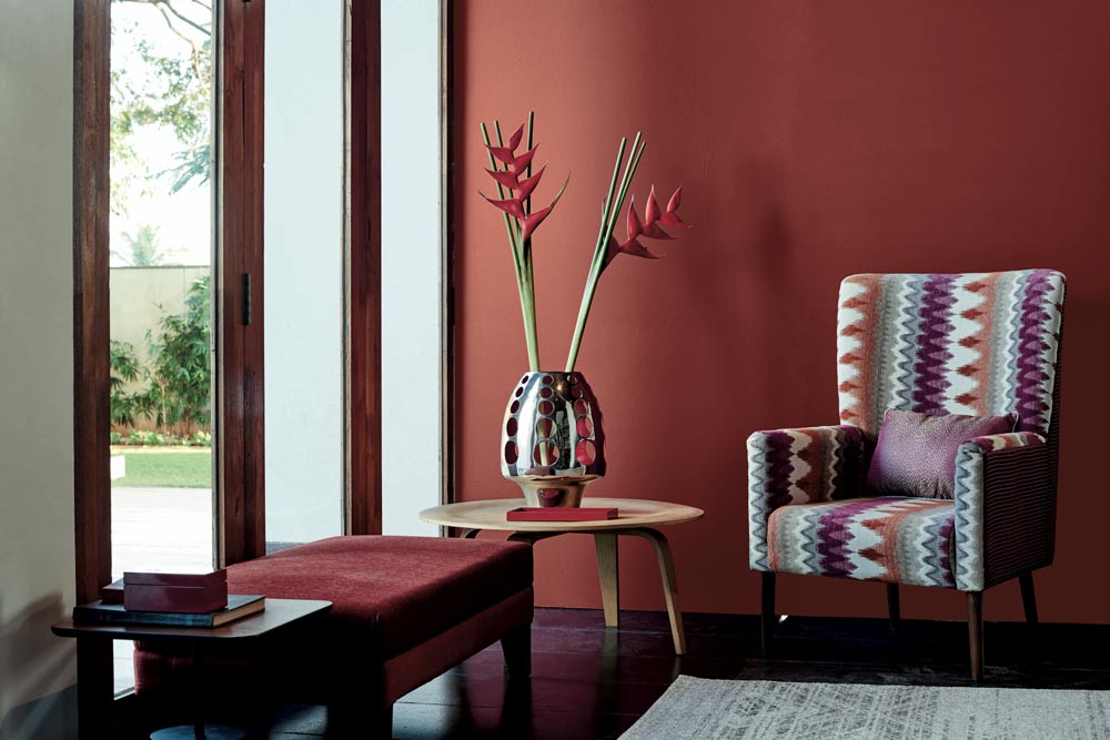 A red room with a chair and a silver vase on a circular wooden table